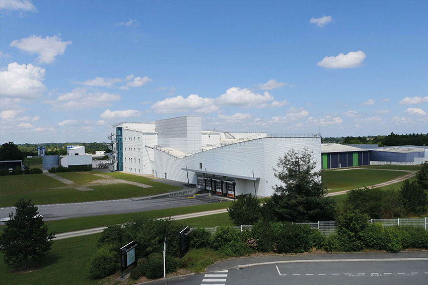 Vue d’un bâtiment industriel blanc entouré d’espaces verts et de routes d’accès.