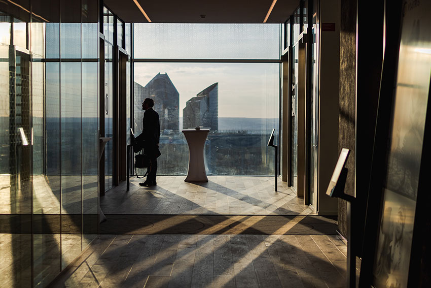 Personne debout près d’une baie vitrée dans un couloir, avec vue sur des immeubles au loin.