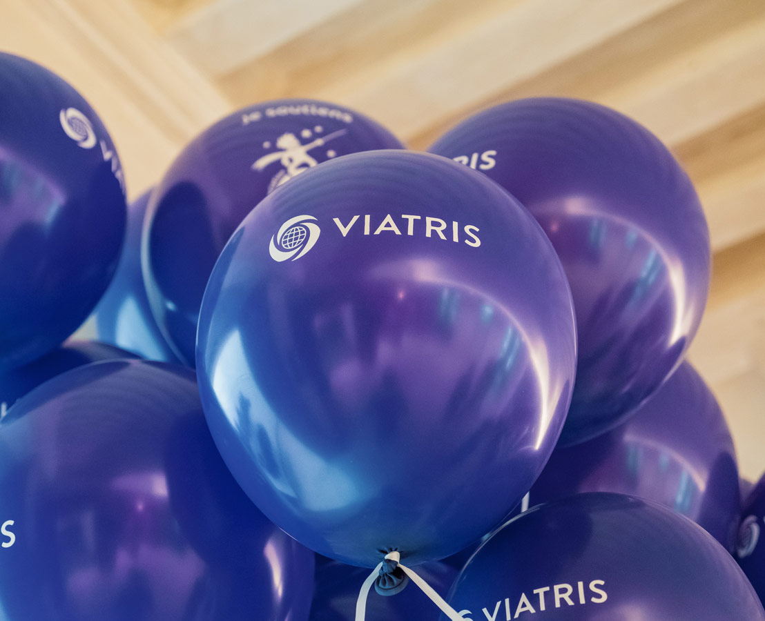 Grappe de ballons violets avec le logo globe et le mot "VIATRIS", flottant sous un plafond en bois dans un espace intérieur.