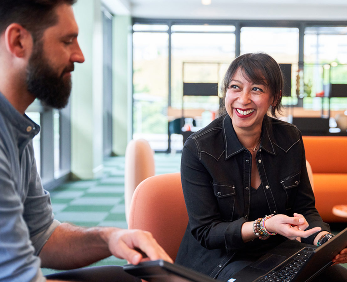 Deux personnes échangent dans un espace lumineux avec de grandes fenêtres ; l’une tient un ordinateur portable, l’autre sourit.