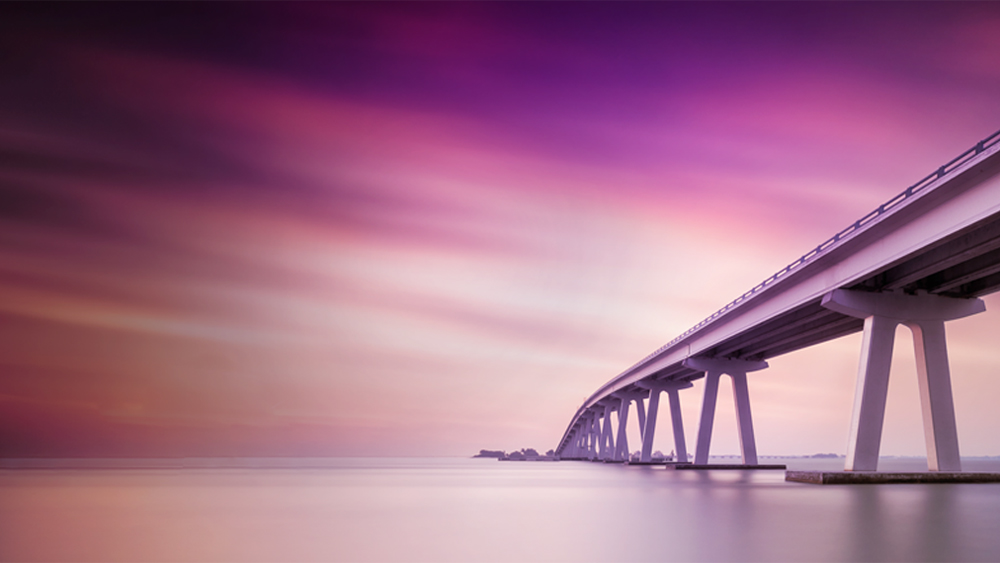 Un long pont moderne s'étendant au-dessus de l'eau au coucher du soleil, dans une ambiance violette et rose.