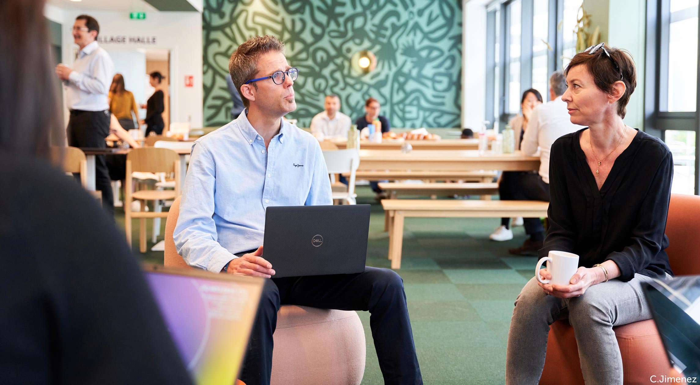 ersonnes assises dans un espace de travail convivial, discutant autour d’ordinateurs et de tasses, avec en arrière-plan une salle lumineuse et décor mural vert