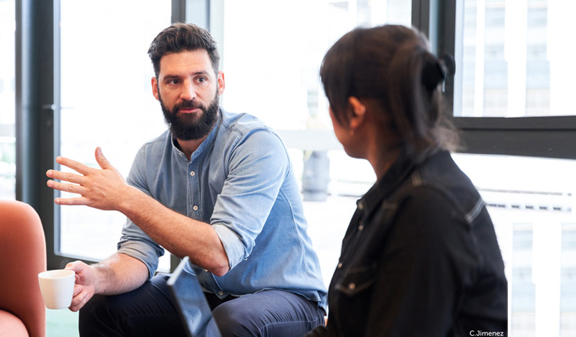 Deux personnes assises dans un espace lumineux, discutant face à face, l’une tenant une tasse, avec de grandes baies vitrées en arrière-plan