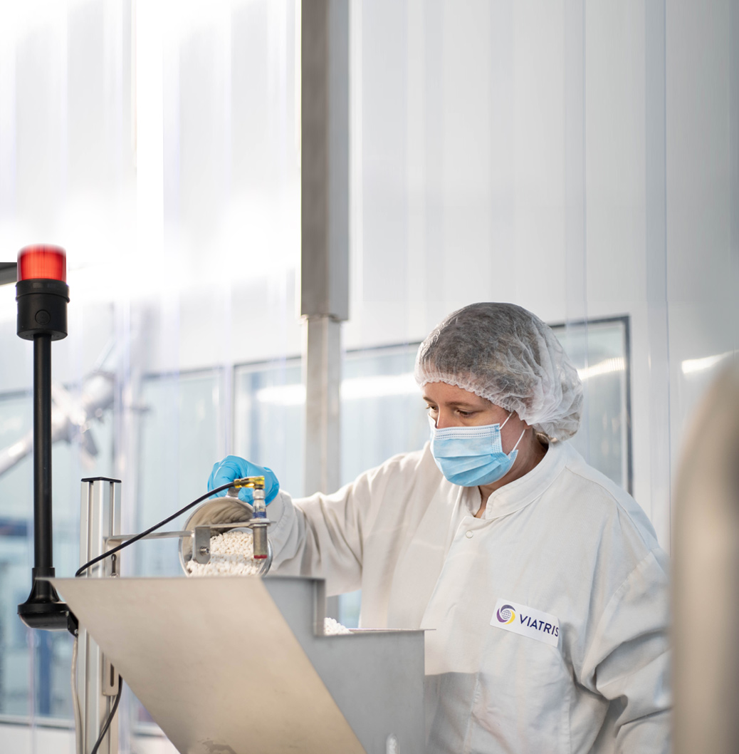 Employée Viatris en blouse de laboratoire et masque chirurgical manipulant des granulés pharmaceutiques dans une salle blanche.