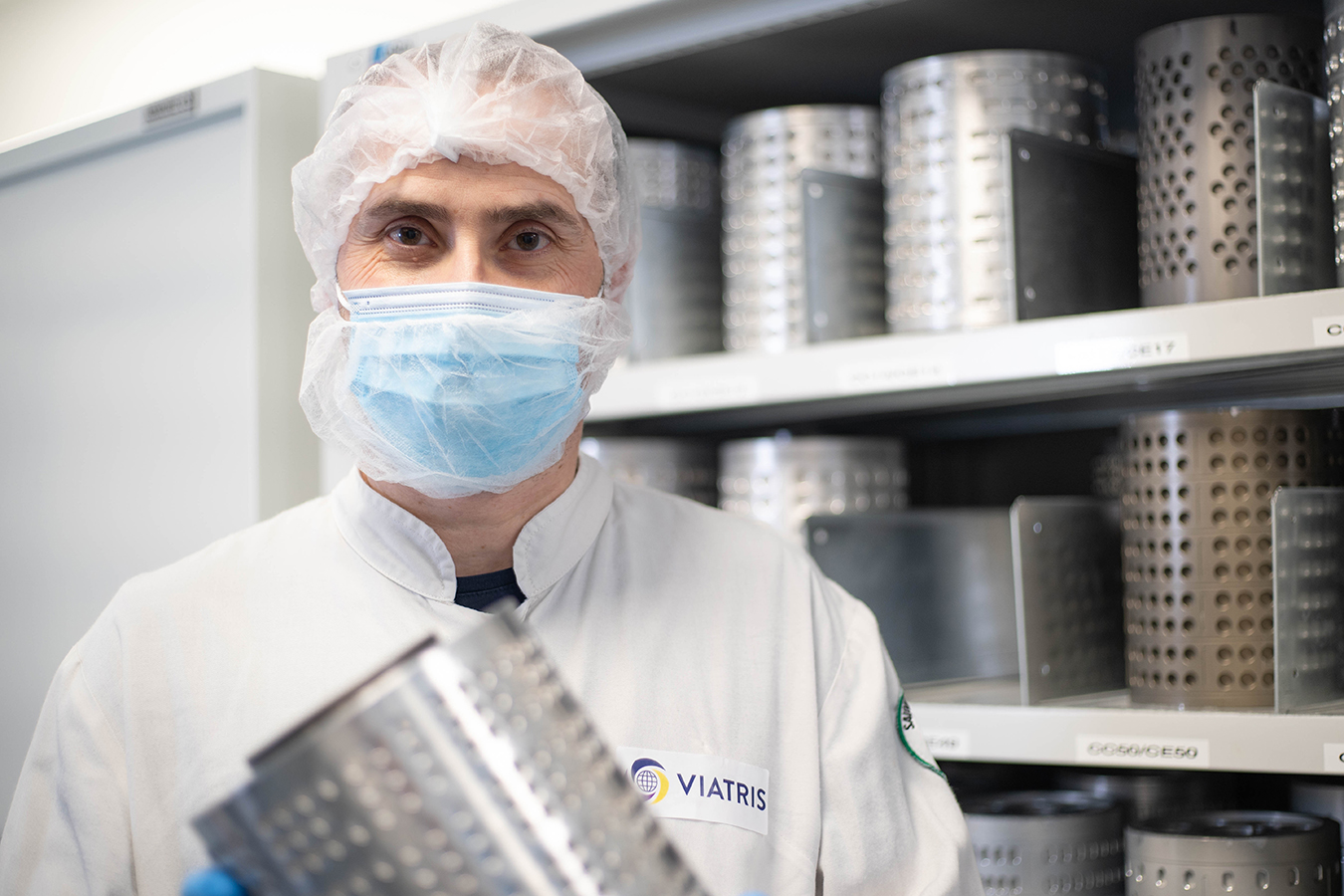 Employé de Viatris en blouse et charlotte tenant un tamis cylindrique, utilisé dans la production pharmaceutique, devant une étagère de pièces similaires
